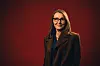 Elisabeth Sørdal med briller i brun frakk står foran rød studiobakgrunn.
