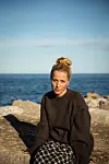 Maria Sand i mørk genser sitter på svaberg foran havet i sollys