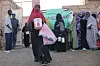 En kvinne bærer matposer og en bøtte utenfor et hjelpekjøkken i Khartoum, mens flere personer står i kø ved en vegg med arabisk skrift.