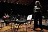 Kristine Tjøgersen står i en konsertsal mens en fiolinist øver blant tomme stoler og notestativer.