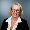Smilende kvinne med briller og lys skjorte foran grå bakgrunn. Portrettfoto Gry Espedal.