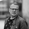 Svart-hvitt-portrett av en mann med briller og jakke mot uskarp bakgrunn.