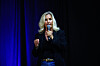 Erin Brockovich speaks into a microphone on a dark stage with blue lighting.