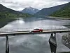 Rød veteranbil på bro over rolig fjellvann med skogkledde åser og snødekte fjell i bakgrunnen.