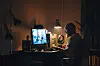 En mann sitter ved et skrivebord i et dunkelt rom og bruker en datamaskin ved en opplyst skjerm.