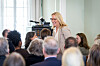 Marit Moum Aune taler foran et publikum i et lyst møterom med kamera og mikrofonstativ.