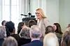 Marit Moum Aune taler foran et publikum i et lyst møterom med kamera og mikrofonstativ.