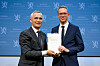 Eigil Knutsen overrekker en rapport til Jens Stoltenberg foran en blå bakgrunn med Stortingets logo.