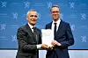Eigil Knutsen overrekker en rapport til Jens Stoltenberg foran en blå bakgrunn med Stortingets logo.