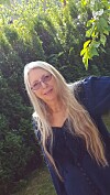 Kvinne med langt blondt hår og briller står foran grønne busker i hagen.