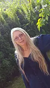 Kvinne med langt blondt hår og briller står foran grønne busker i hagen.