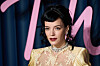 Lily Allen står foran en mørk bakgrunn med rosa skrift, iført lys gul kjole og smykker.