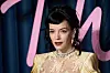 Lily Allen står foran en mørk bakgrunn med rosa skrift, iført lys gul kjole og smykker.