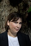 Portrett av en person foran en grov trestamme, med svart blazer og hvit topp.