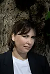 Portrett av en person foran en grov trestamme, med svart blazer og hvit topp.
