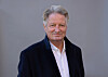 Mann i mørk jakke og hvit skjorte foran grå bakgrunn.