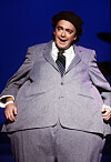 Øivind Blunck står på scenen i grå dress og svart hatt foran blå bakgrunn.