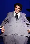 Øivind Blunck står på scenen i grå dress og svart hatt foran blå bakgrunn.