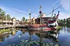 Piratskip ved brygge i Dyreparken i Kristiansand, omgitt av vann, trær og refleksjoner.