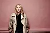 Margreth Olin står foran en rosa vegg i lys beige jakke.