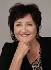Smilende Anne B. Ragde med mørkt hår og svart jakke mot lys bakgrunn.