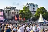 Stor folkemengde på Palmesus ved Bystranda i Kristiansand, med scene, trær, bygninger og seilbåt i bakgrunnen.