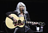 Emmylou Harris synger i mikrofon og spiller akustisk gitar på en mørklagt scene.