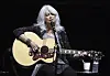 Emmylou Harris synger i mikrofon og spiller akustisk gitar på en mørklagt scene.