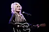 Dolly Parton synger i mikrofonen og spiller gitar på en mørklagt scene.