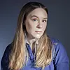 Ung kvinne med langt lyst hår og blå skjorte fotografert mot mørk bakgrunn.