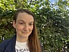 Smilende kvinne i mørk jakke foran frodig grønt løvverk.
