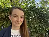 Smilende kvinne i mørk jakke foran frodig grønt løvverk.