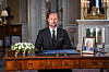 Kong Haakon sitter bak et utsmykket skrivebord i Slottet med blomster og portrett av Kong Harald.