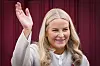 Kronprinsesse Mette-Marit smiler og vinker foran en mørkerød bakgrunn.
