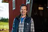 Jon Almaas smiler foran en rød låve på gården sin, med åkerlandskap i bakgrunnen.