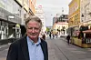 Eldre mann i mørk jakke står på en travel gågate med butikker og bygninger bak seg.