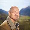 Portrett av en smilende, skjeggete mann i jakke foran et fjellandskap.