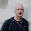 Nærportrett av en mann i mørk genser foran en lys murvegg.
