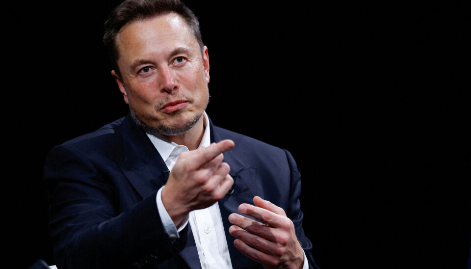 Elon Musk i mørk dress gestikulerer mot publikum foran en mørk bakgrunn.