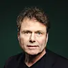 Nærbilde av en mann med brunt hår i mørk jakke mot grønn bakgrunn.