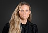 Kvinne med langt, bølgete blondt hår og svarte klær foran en mørk bakgrunn.