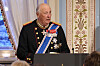 Kong Harald i uniform holder tale ved en talerstol i en rikt dekorert sal.