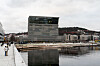 Munchmuseet og speilbildet i vannet sett fra promenaden i Oslo, med mennesker langs kaikanten.