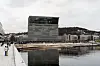 Munchmuseet og speilbildet i vannet sett fra promenaden i Oslo, med mennesker langs kaikanten.