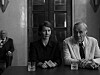 Kvinne og eldre mann sitter ved et bord i et dunkelt rom, med en person i bakgrunnen.