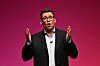 Andy Burnham taler med hendene hevet foran en magentarød bakgrunn.