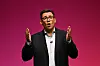 Andy Burnham taler med hendene hevet foran en magentarød bakgrunn.