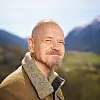 Skjeggete mann i brun jakke smiler foran fjell, skog og et grønt dalføre.