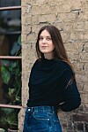 Siv Helén Strømland i svart topp og blå jeans foran en gammel murvegg og et vindu med planter.