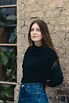 Siv Helén Strømland i svart topp og blå jeans foran en gammel murvegg og et vindu med planter.